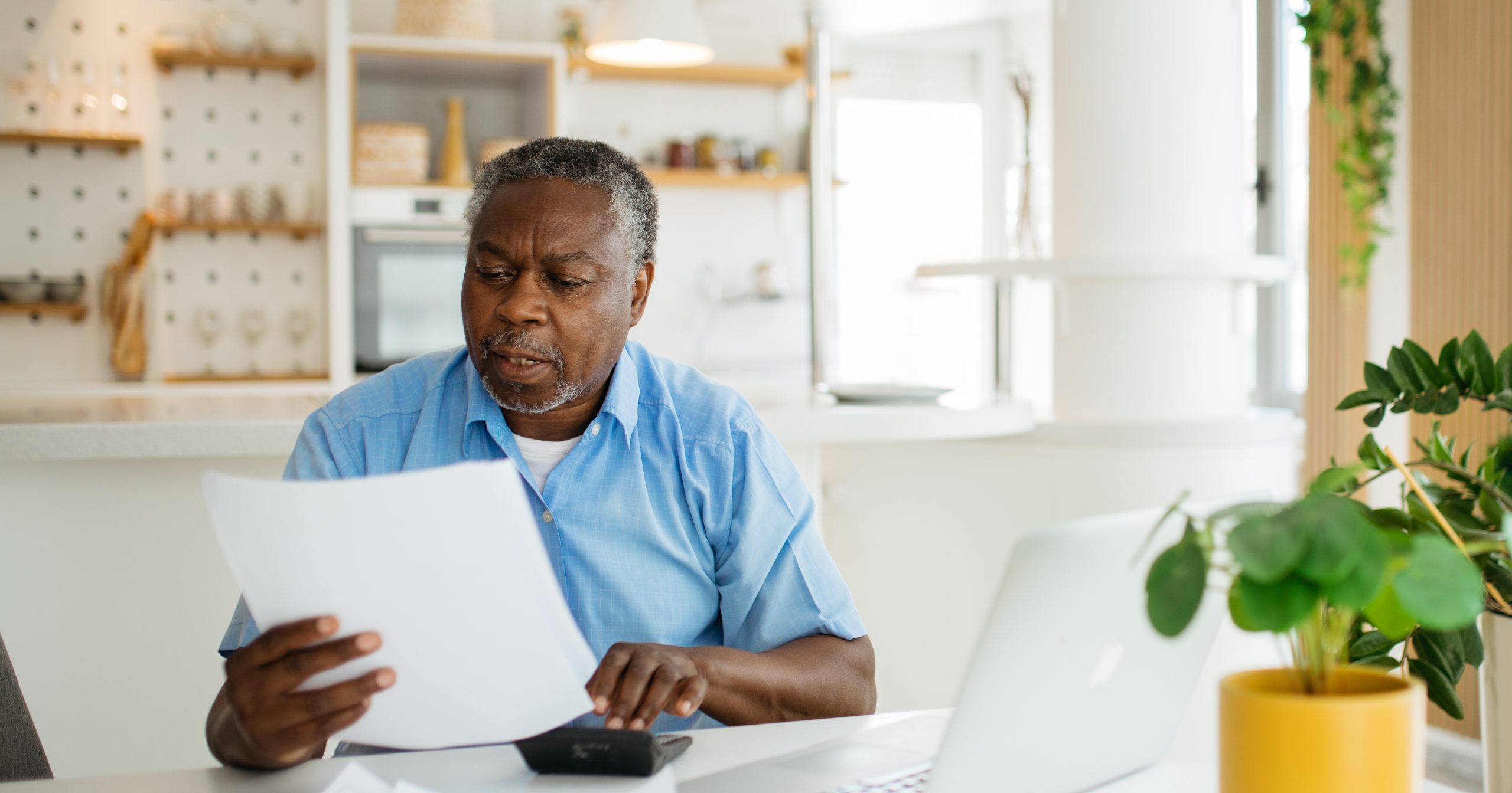 A man sitting behind a laptop with his calculator looks at a sheet of paper