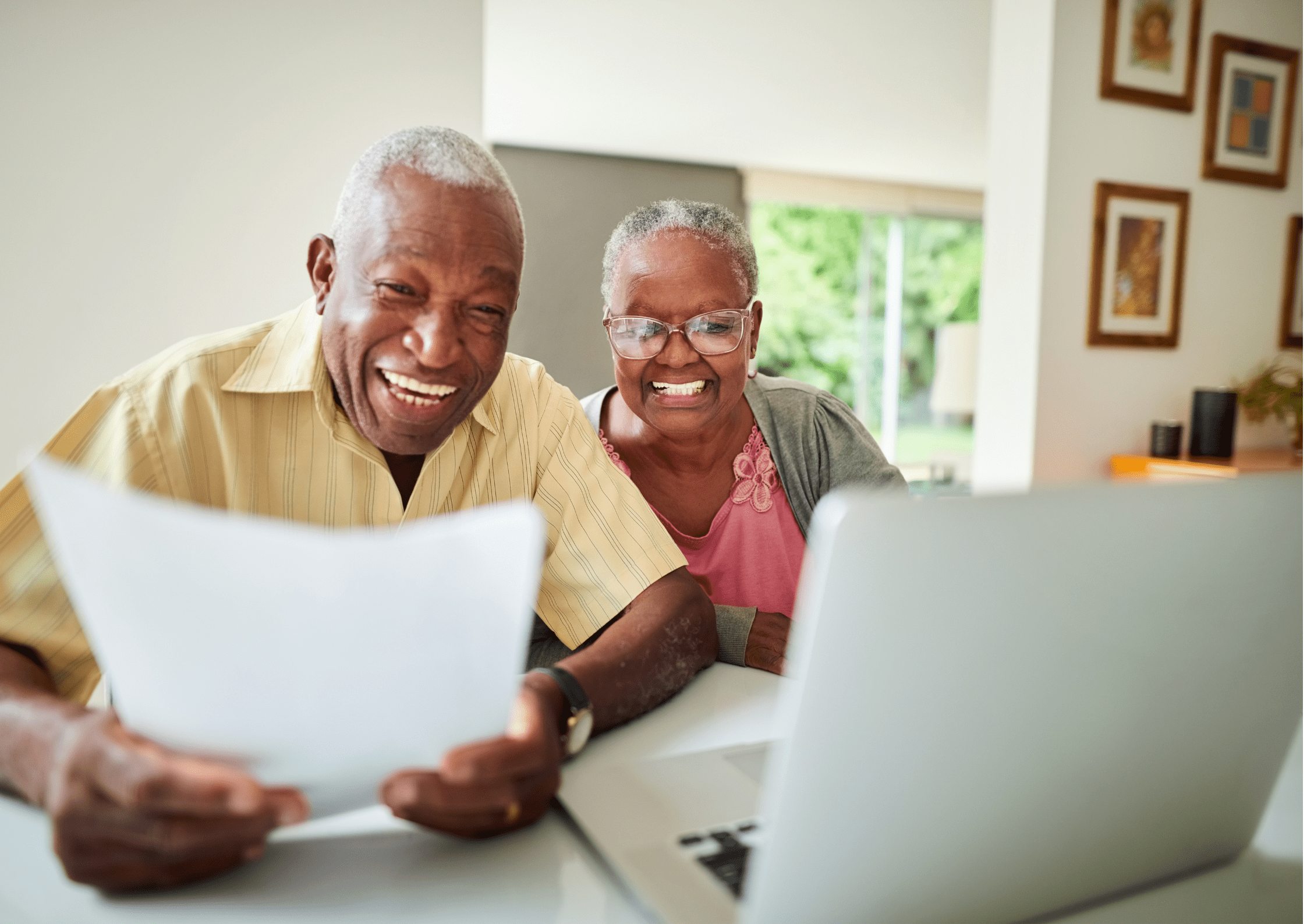 Older couple is looking at a laptop while smiling and the man holds a paper in his hand