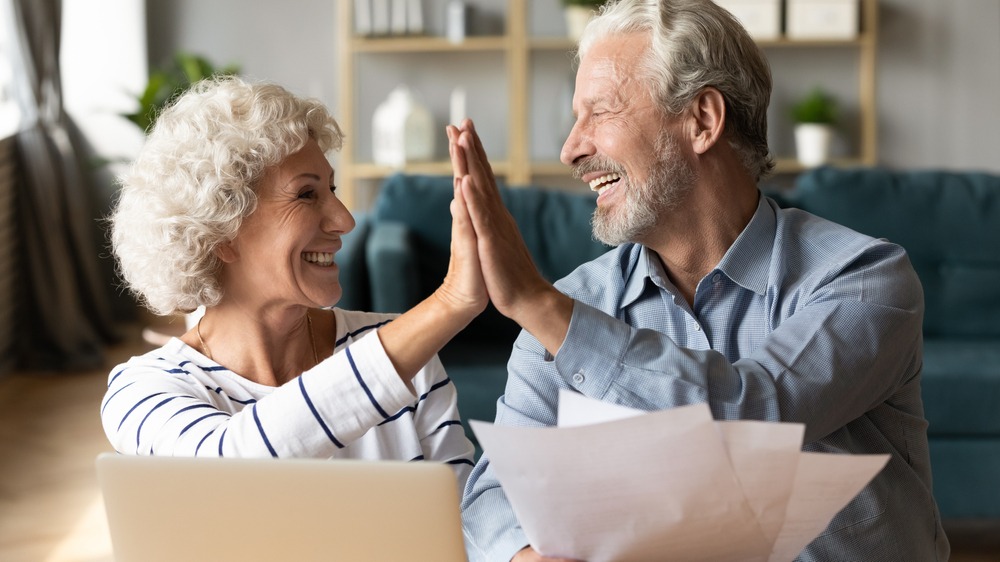 Older couple gives each other a high five while laughing at each other. The man holds some papers in his hand and there is a laptop in front of them